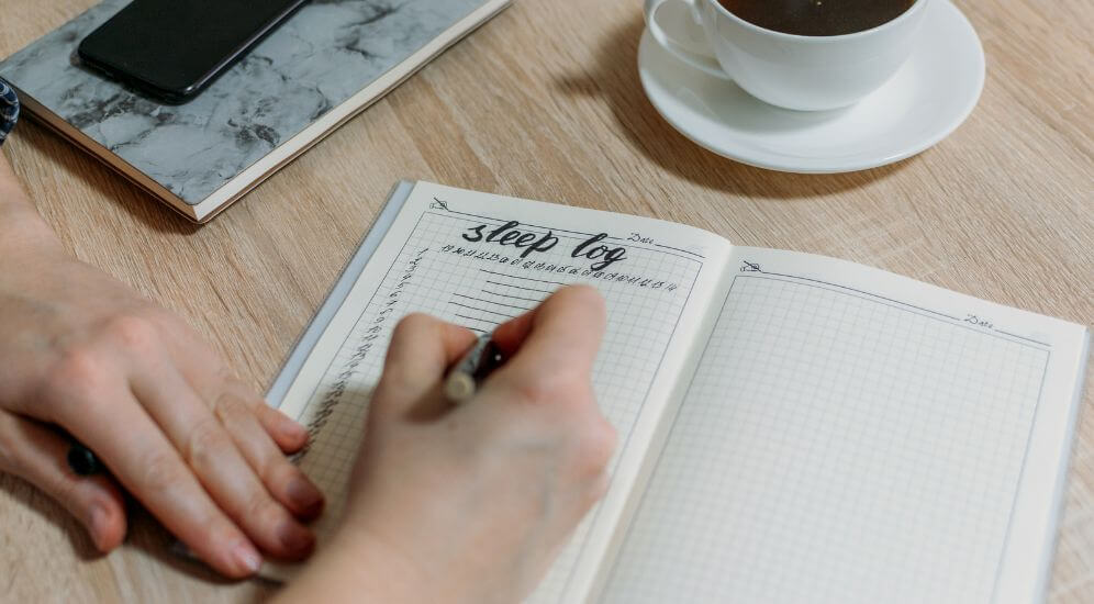 A bedside table with a notebook open to a handwritten sleep diary next to a pen and a softly glowing lamp in a quiet bedroom