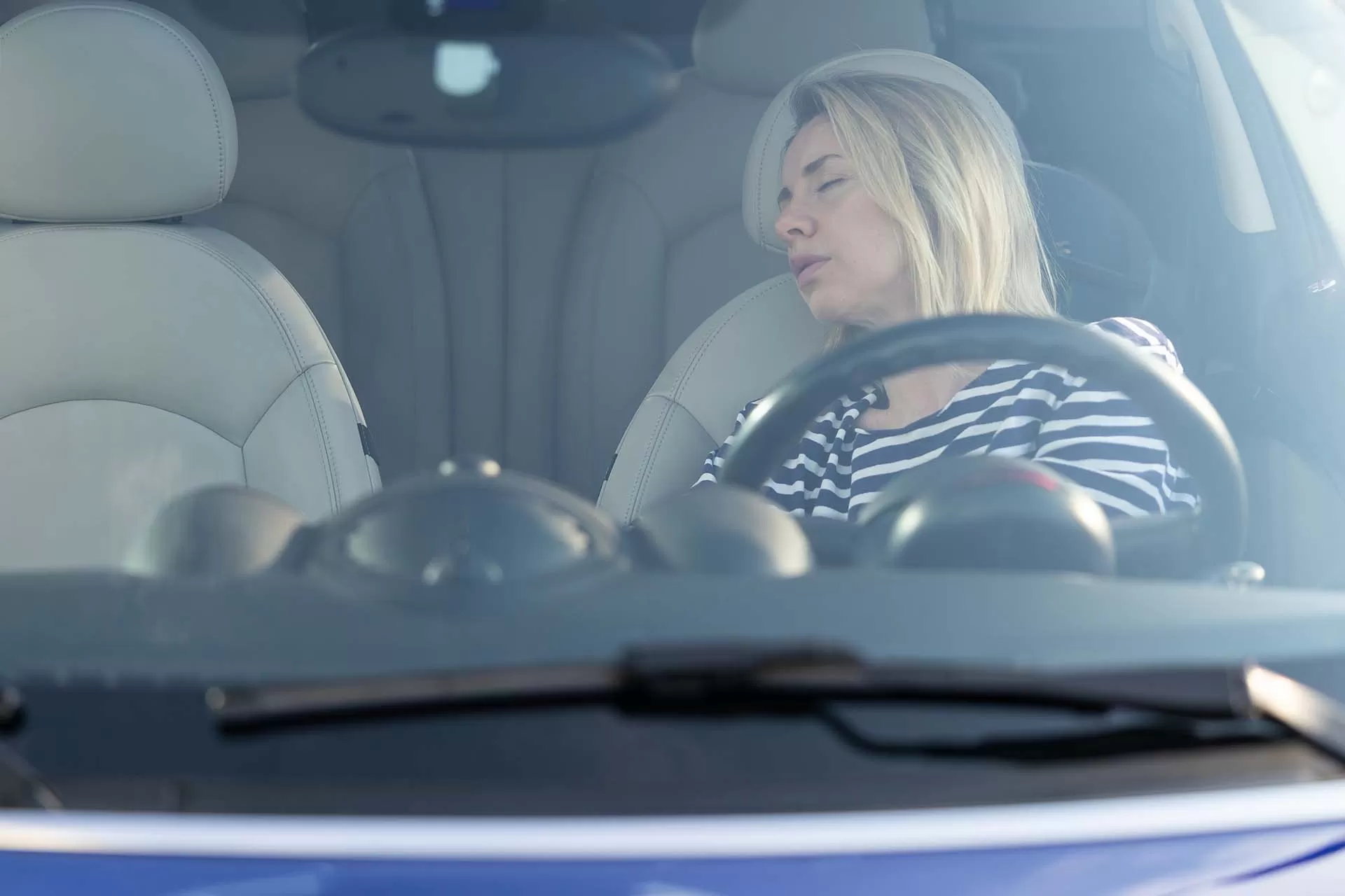 A driver in a parked car with both hands on the steering wheel, eyes heavy and posture slumped as if struggling to stay awake, natural light through the windshield