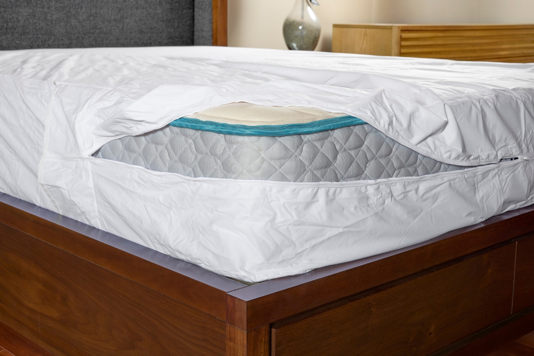 A mattress encasement being zipped fully closed around a mattress, with the zipper pull and fabric seam clearly visible in bedroom lighting