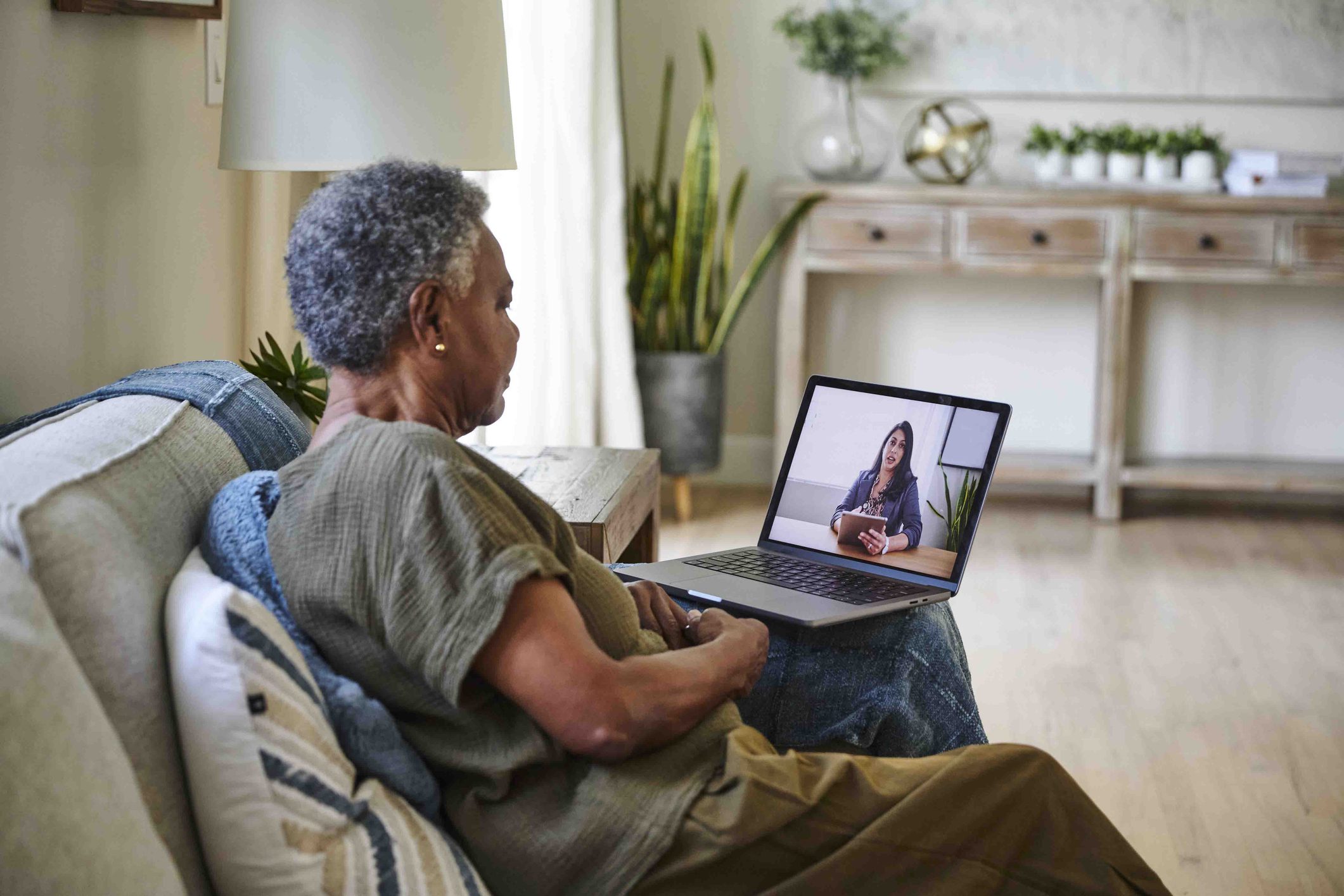A person seated at a kitchen table attending a telehealth therapy session on a laptop, with a notebook and a warm drink nearby in soft morning light