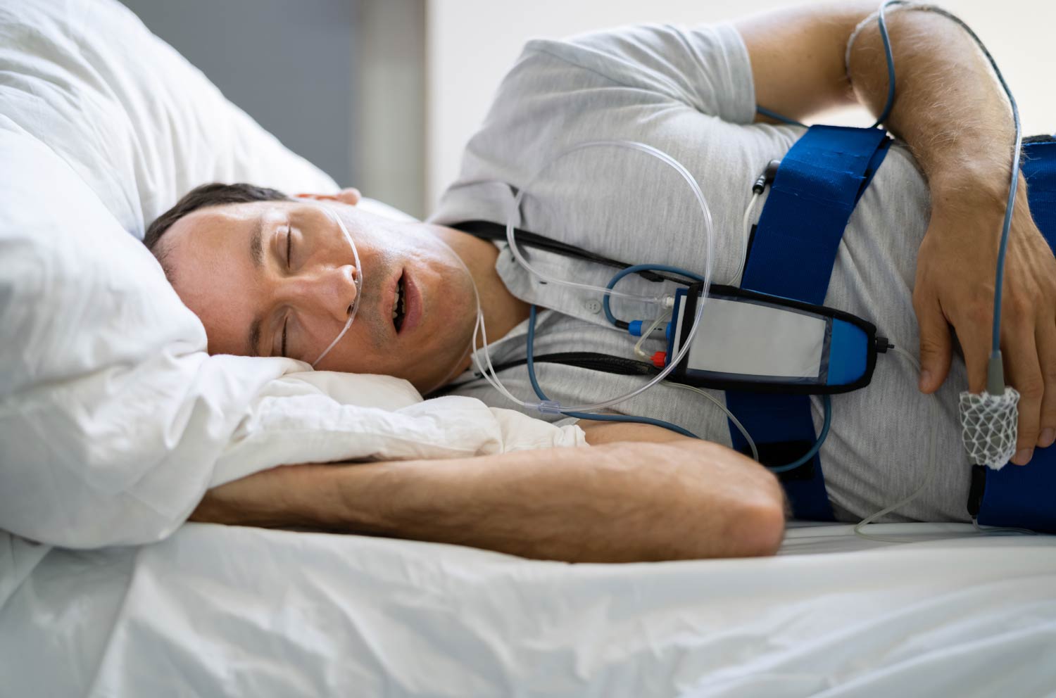 An adult patient sitting on the edge of a bed adjusting a nasal cannula and a finger pulse oximeter for a home sleep apnea test