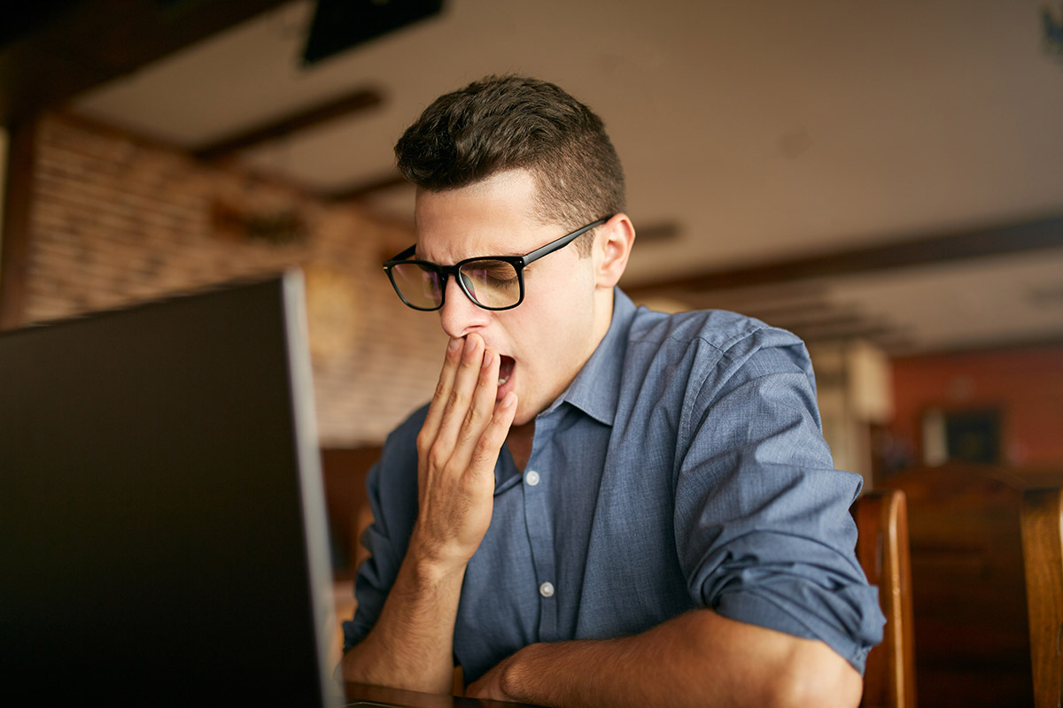 An adult sitting at an office desk in afternoon light, yawning with eyes partly closed while a laptop and notebook sit open in front of them